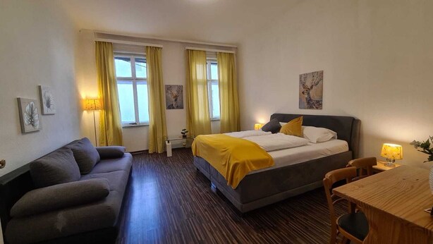 A hotel room with a bed, a sofa, a table, and two windows with curtains. - Cozy 1BR Apt Long Stay for 3 Guests (Vienna)