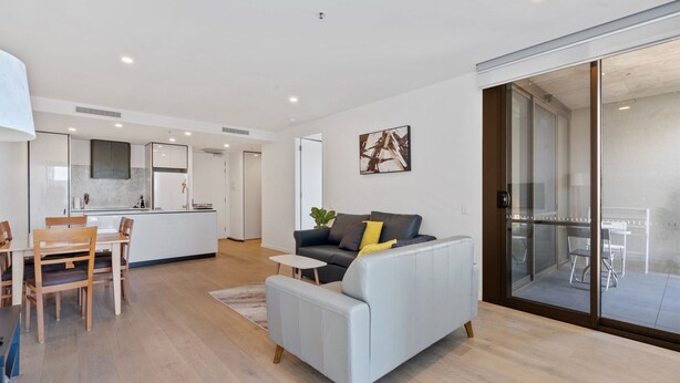 Een moderne woonkamer met een grijze bank, een eethoek met houten stoelen, een keuken achtergrond en een schuifpui naar een balkon.
