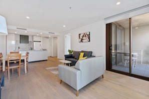 Un salon moderne avec un canapé gris, un coin repas avec des chaises en bois, une cuisine en arrière-plan et une porte-fenêtre coulissante donnant sur un balcon.