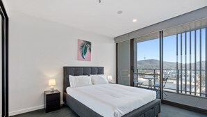 Een moderne hotelkamer met een groot bed, een nachtkastje met een lamp en een glazen deur die uitkomt op een balkon met uitzicht op de stad.