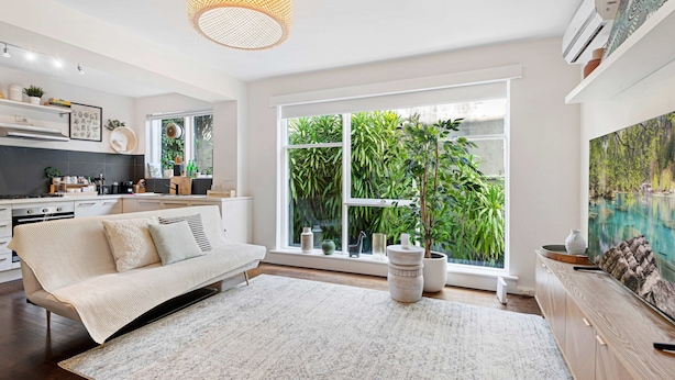 A modern kitchen with white cabinetry, a dining area with a white sofa, and a large window with a view of greenery. - Contemporary 1-bed Unit Near Chapel St (Melbourne)