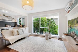 A modern kitchen with white cabinetry, a dining area with a white sofa, and a large window with a view of greenery. - Contemporary 1-bed Unit Near Chapel St (Melbourne)