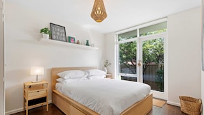 A modern bedroom with a wooden bed, a bedside table, a nightstand, a hanging light fixture, and a large window with a view of greenery outside. - Contemporary 1-bed Unit Near Chapel St (Melbourne)