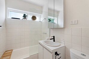A modern bathroom with white tiles, a white sink, a mirror, and a window above the sink.