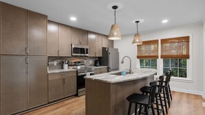 A modern kitchen with wooden cabinets, stainless steel appliances, a central island with bar stools, and a window with blinds. - Spacious|Close to DT|5 BDRM|4BA|PetsOK (Greenville)