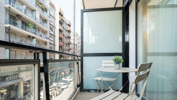 A balcony with a small table and chairs, a potted plant, and a view of a busy street with cars and buildings. - Barirooms - District 198 (Bari)
