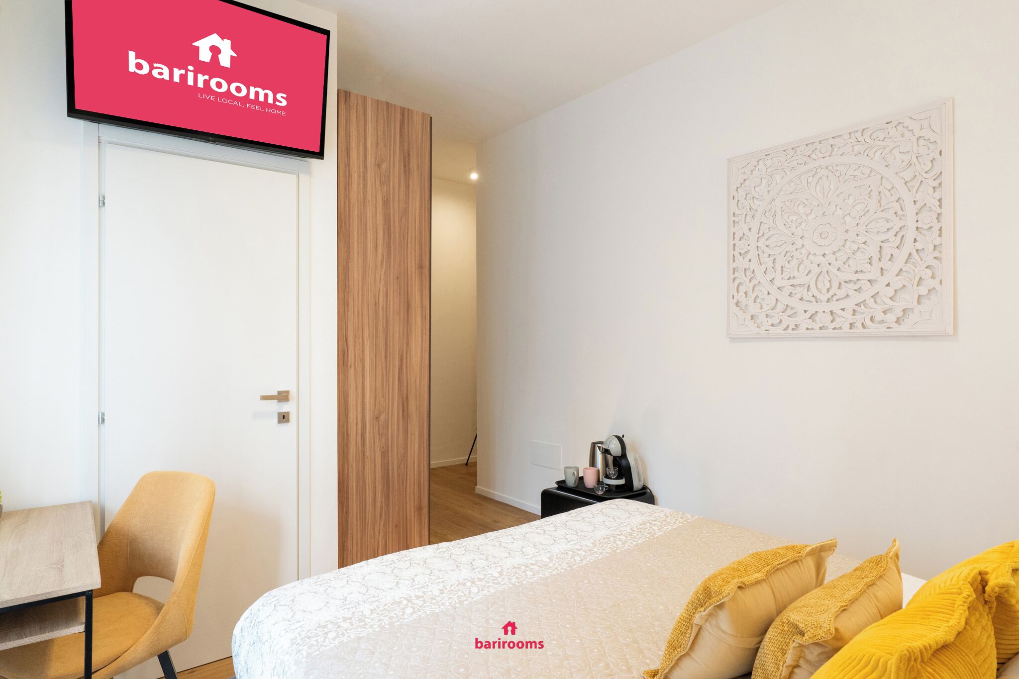 A hotel room with a bed, a chair, a desk, a coffee machine, and a TV displaying the barrirooms logo.
