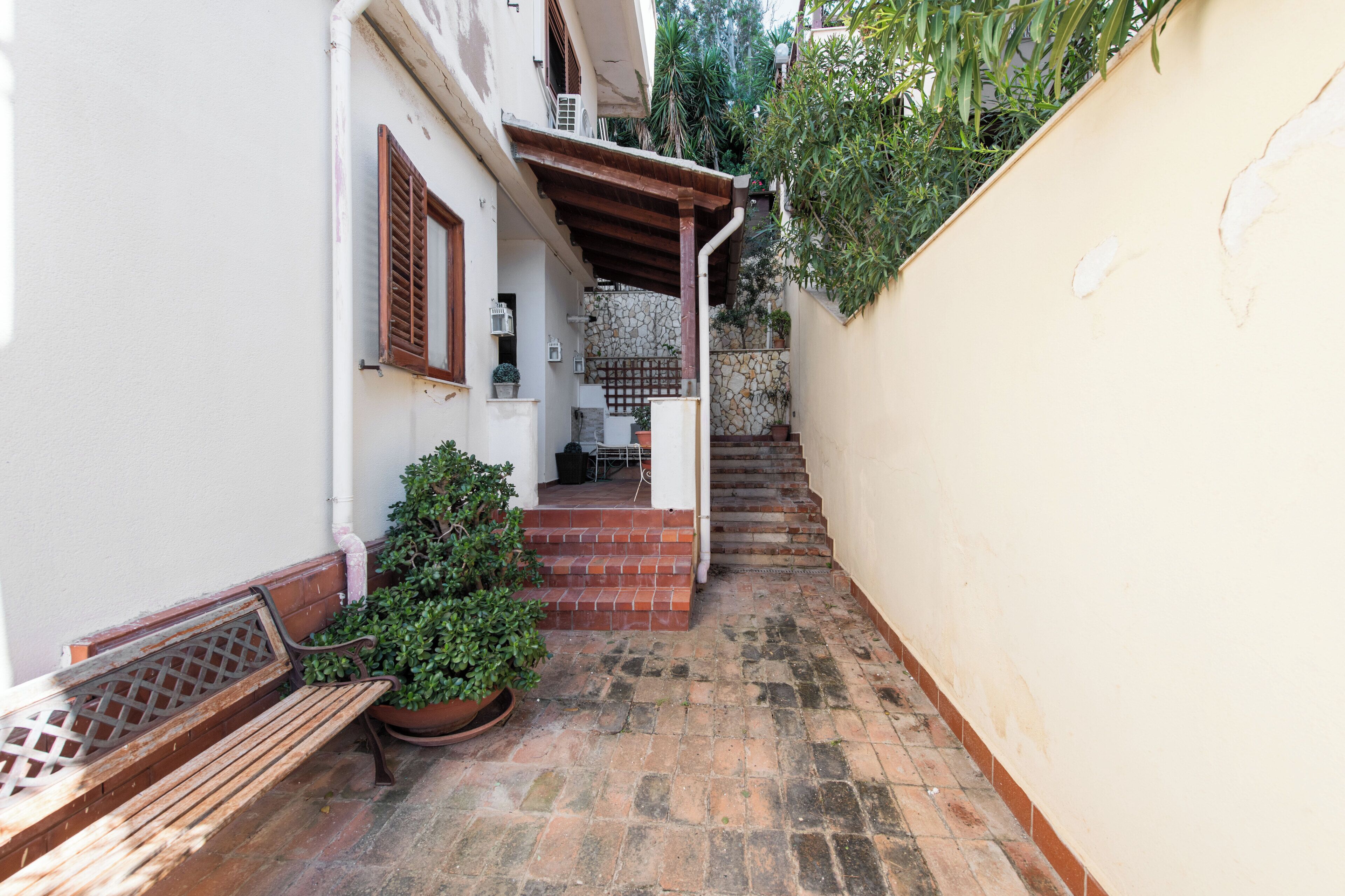 A narrow alleyway with a wooden bench, potted plants, and a staircase leading to a stone wall.