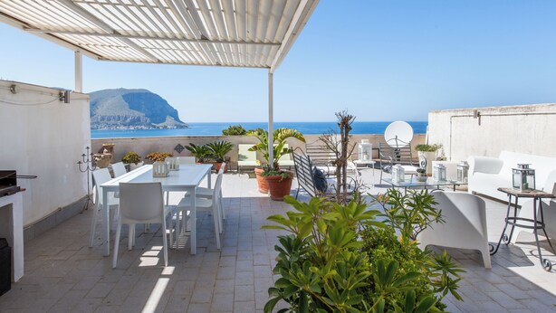 Une terrasse avec une table blanche, des chaises et des plantes en pot, d’où l’on peut admirer une mer bleue et limpide et une montagne.