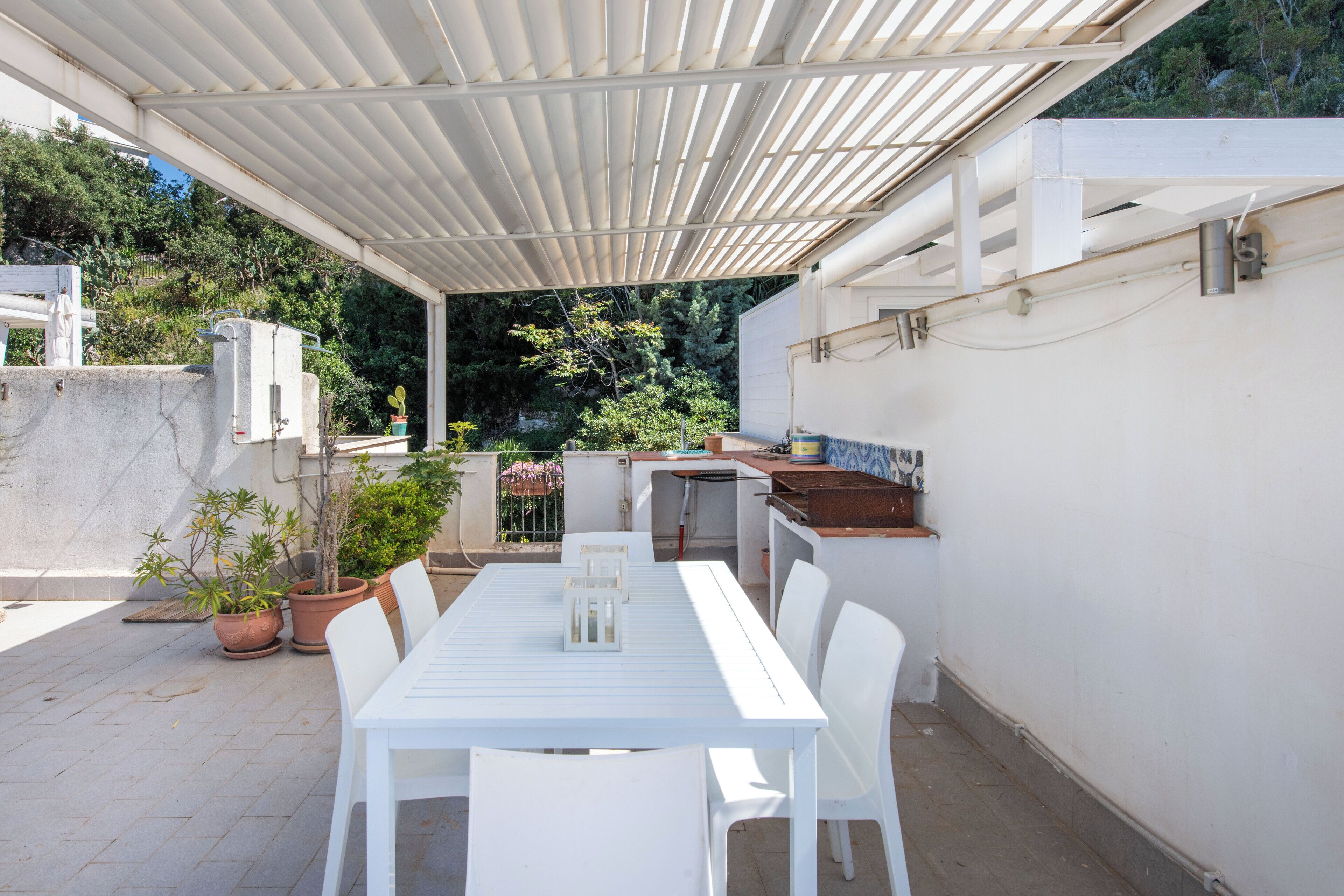 Een terras buiten met een witte tafel en stoelen, omringd door potplanten en een barbecuehoek.