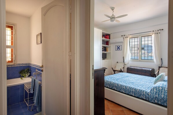 A bedroom with a bed, a ceiling fan, a window with curtains, and a small shelf with books.
