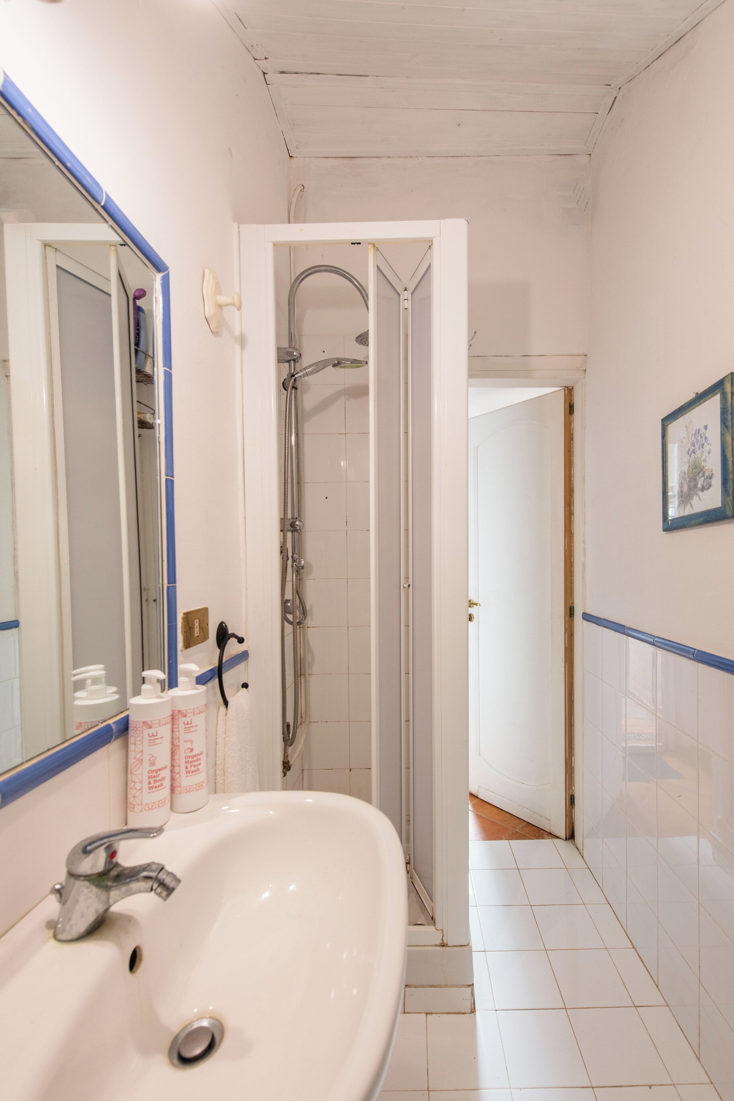 Een badkamer met een witte wastafel, een spiegel, een douche en een deur naar een andere kamer.