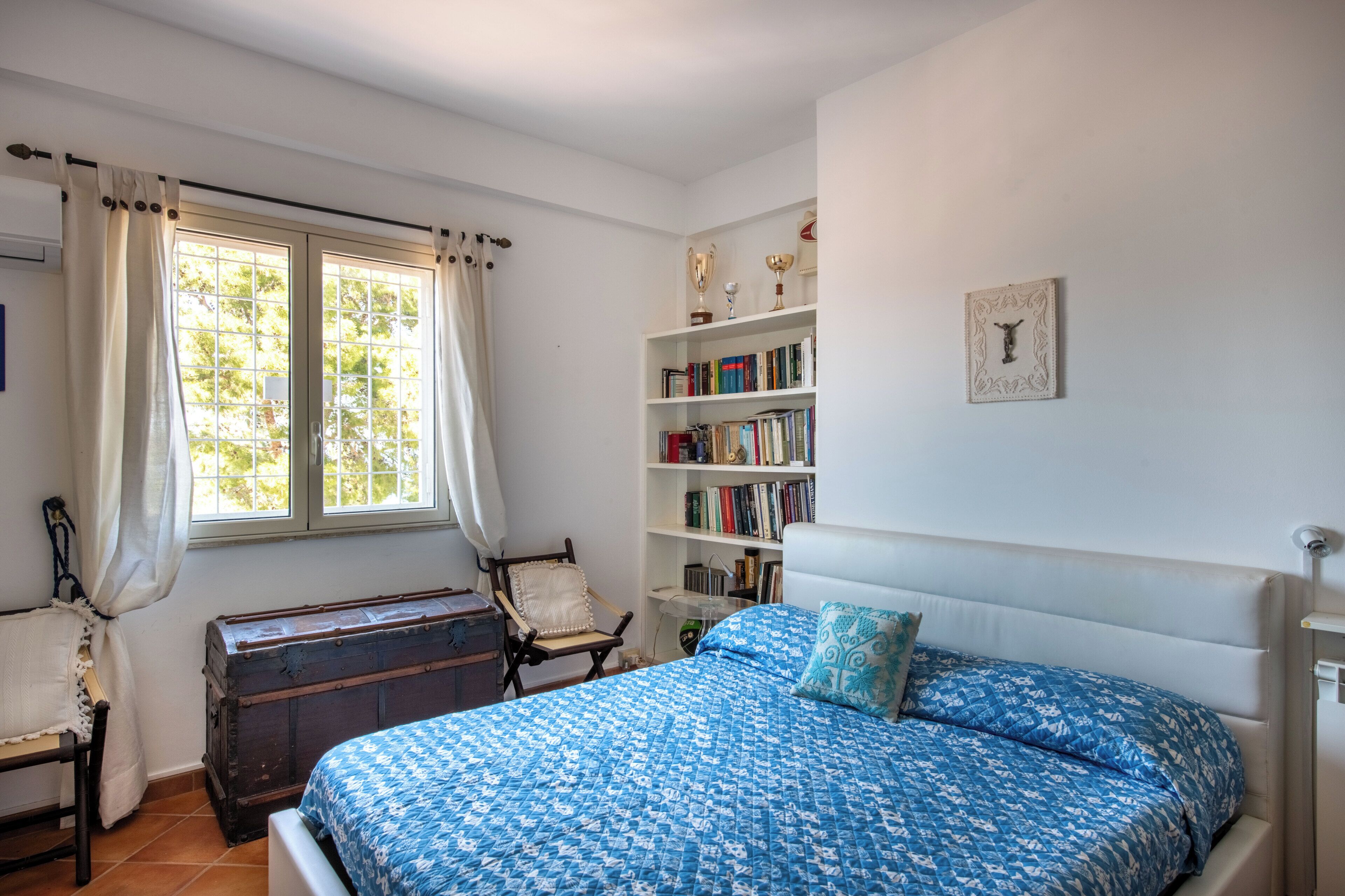 A bedroom with a bed, a bookshelf, a chair, a window with curtains, and a wooden chest.