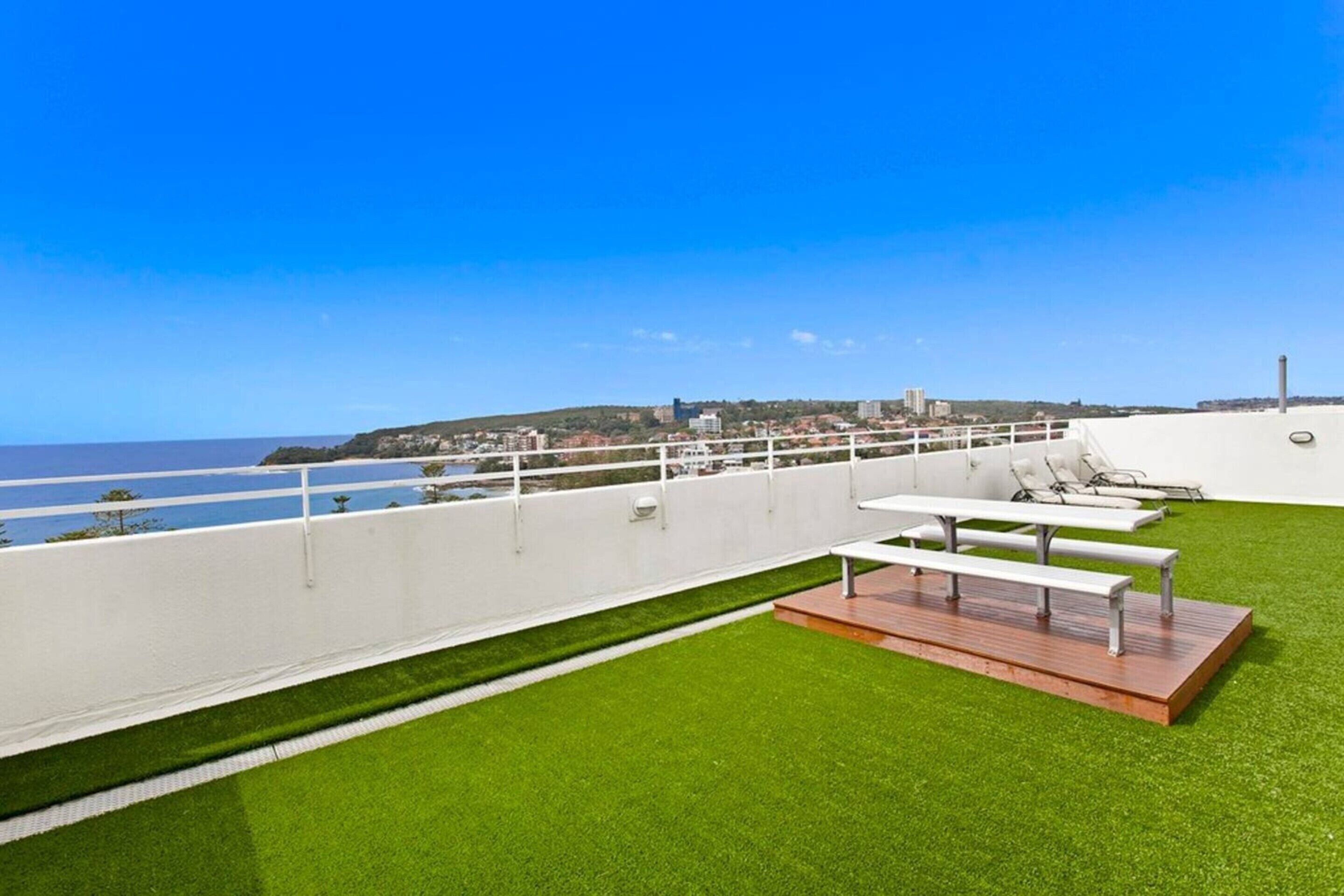 Una terraza en la azotea con césped artificial, barandillas blancas y vistas al mar y edificios a lo lejos.