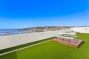 Una terraza en la azotea con césped artificial, barandillas blancas y vistas al mar y edificios a lo lejos.