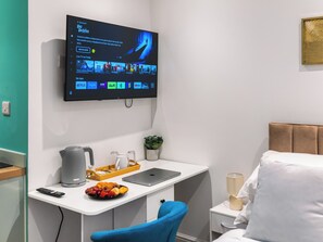 Habitación de hotel con televisión de pantalla plana montada en la pared mostrando una película, un escritorio con una computadora portátil, una silla, una mesita de noche con lámpara y una cama con ropa blanca de cama.