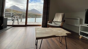 Une pièce dotée d’un plancher en bois, d’une chaise, d’une table, d’un radiateur et offrant une vue sur une montagne et un lac, grâce à de grandes portes coulissantes.