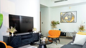 A modern living room with a large flat-screen TV, a sofa, an orange chair, and a coffee table with decorative items. - Charming 2-bedroom apartment with AC in vibrant Dubai-Jumeira Village Circle (Dubai)