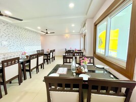 Un espace repas comprenant plusieurs tables et chaises, des ventilateurs de plafond et de grandes fenêtres donnant sur l’extérieur.