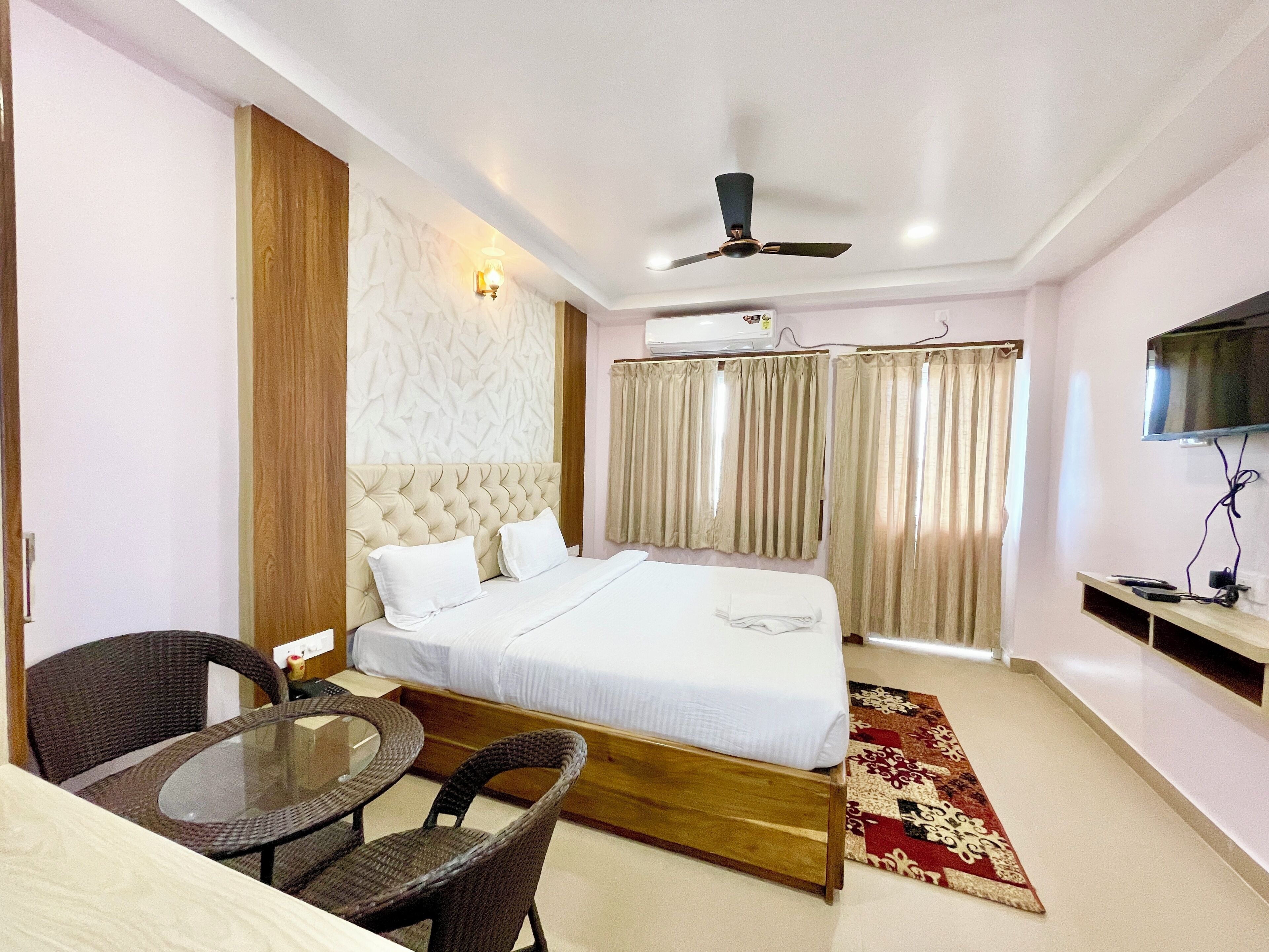 A hotel room with a bed, a ceiling fan, a TV, a table with chairs, and a patterned rug.