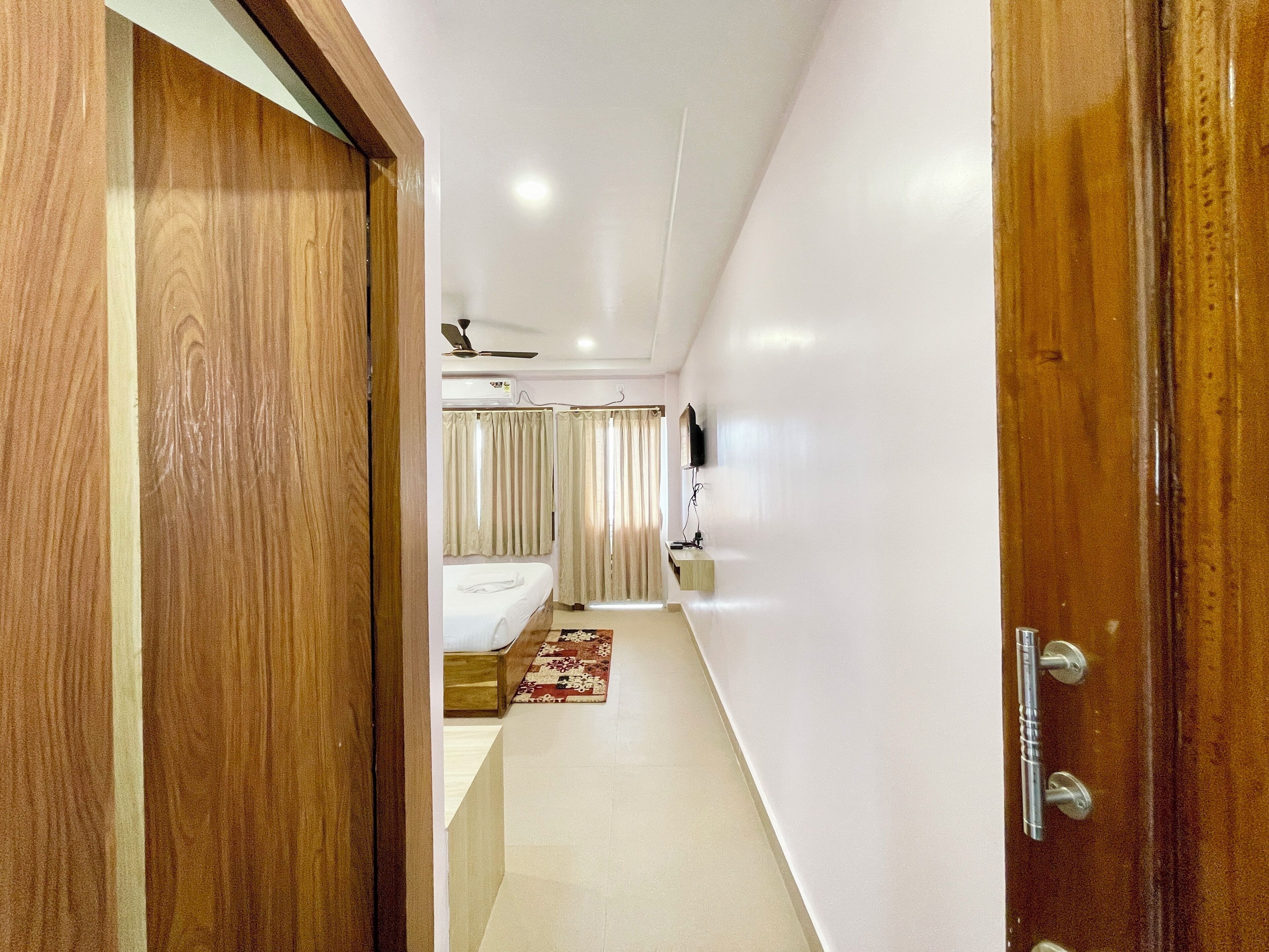 A narrow hallway with a wooden door on the left, a white wall, and a small bathroom visible in the distance with a toilet, sink, and shower curtain.