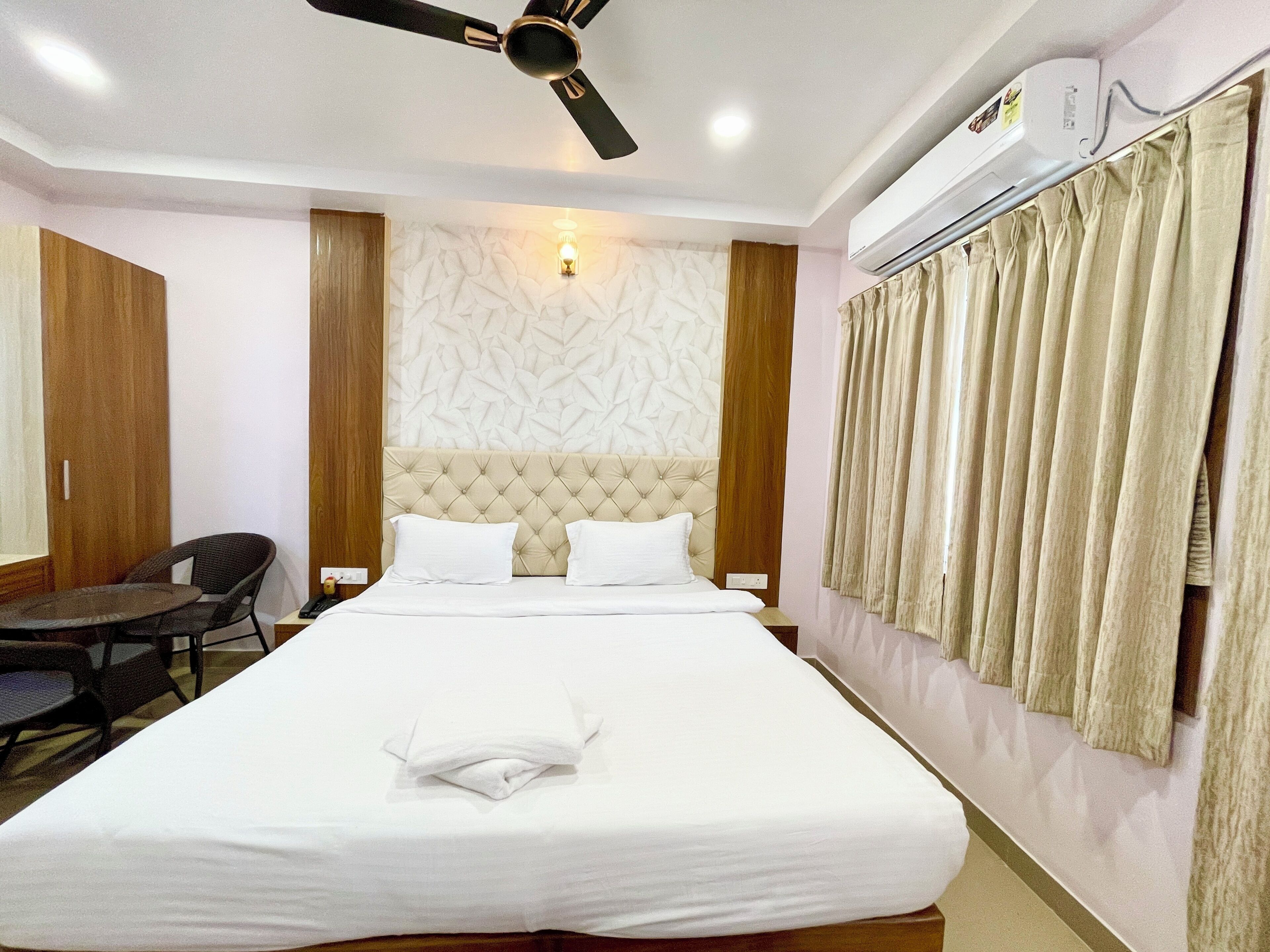 Une chambre d’hôtel avec un grand lit, un ventilateur de plafond, un appareil de climatisation, une lampe fixée au mur et une fenêtre avec des rideaux.