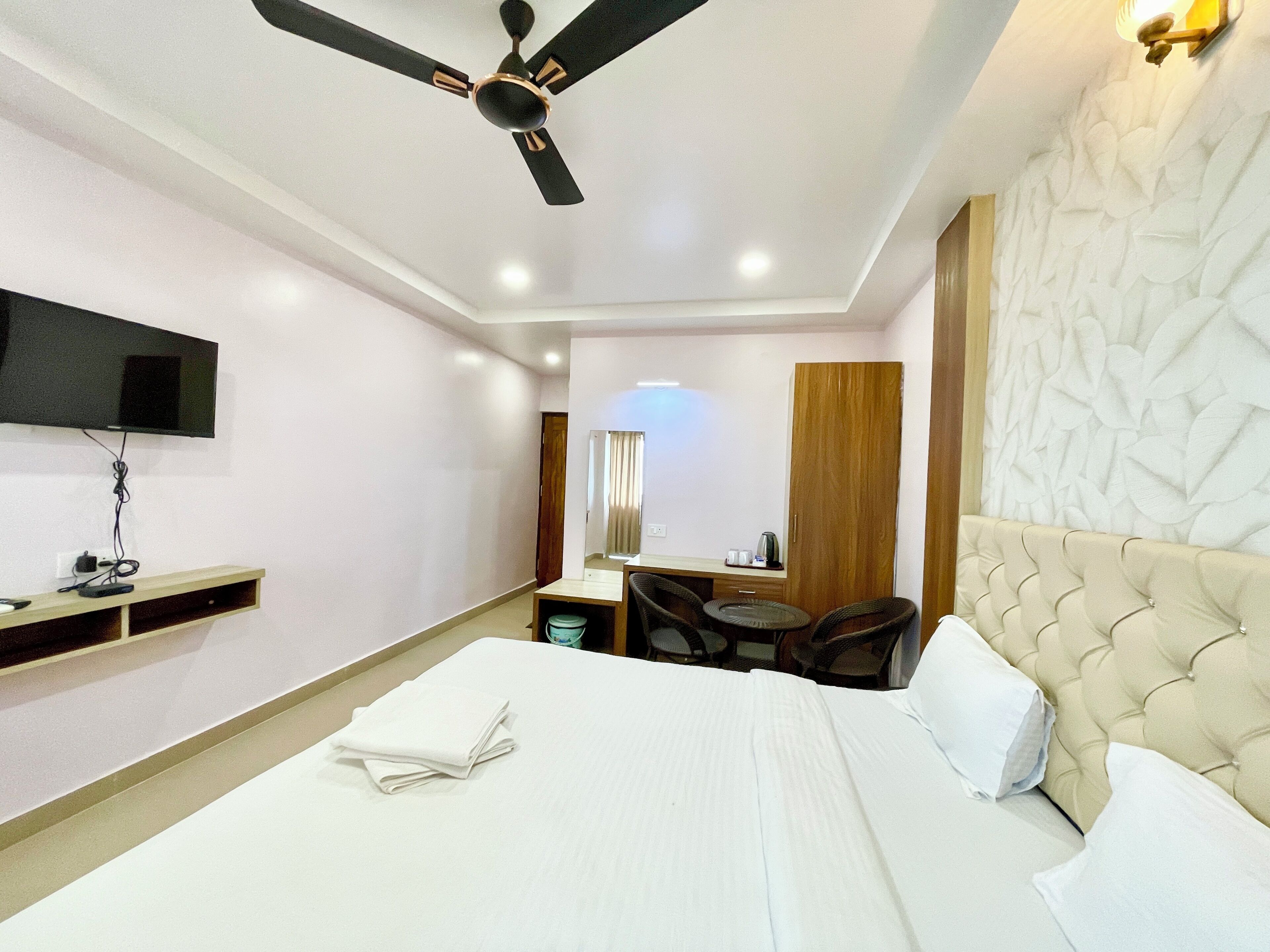 A hotel room with a bed, a ceiling fan, a TV mounted on the wall, a small table with chairs, and a wooden wardrobe.