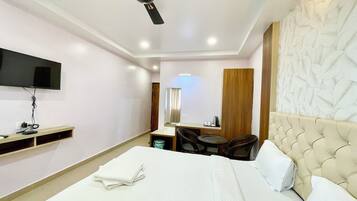 Quarto de hotel com cama, ventilador de teto, televisão fixada na parede, mesa pequena com cadeiras e guarda-roupa de madeira.