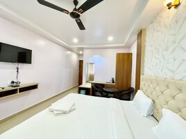 Quarto de hotel com cama, ventilador de teto, televisão fixada na parede, mesa pequena com cadeiras e guarda-roupa de madeira.