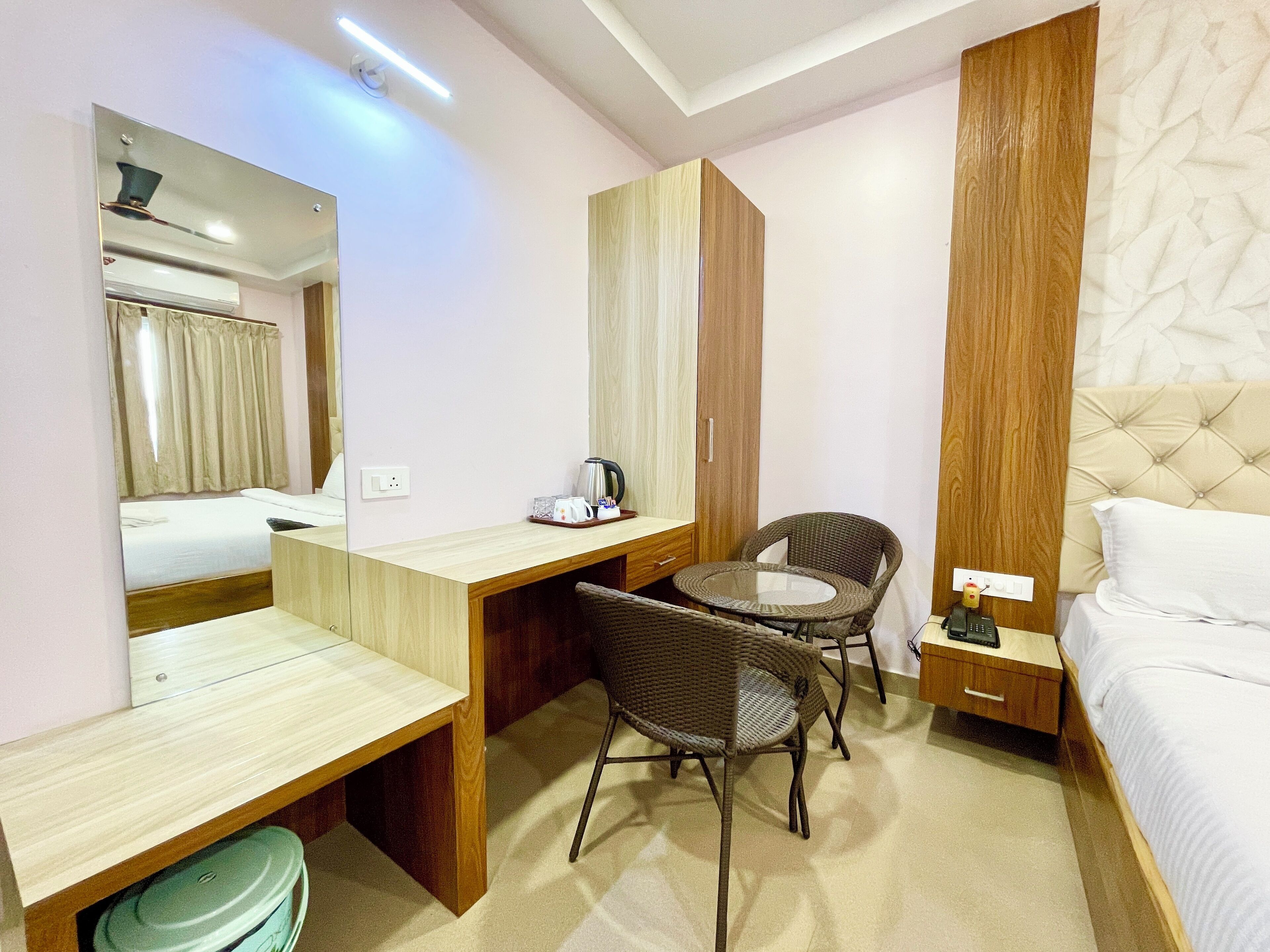 A hotel room with a bed, a desk with a chair, a small table, a mirror, and a wardrobe.