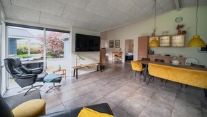 Een moderne woonkamer met een groot raam, een flatscreen televisie, een eethoek met gele stoelen en een keuken met houten kastjes.