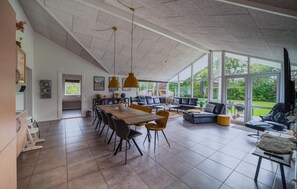 A spacious living area with a large dining table, sofas, and a view of greenery outside.