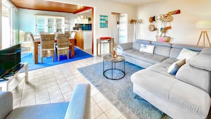 A living room with a grey sofa, a dining area with wicker chairs, and a kitchen in the background.