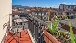 Un balcone con un tavolo rotondo, piante in vaso e vista su una zona residenziale con edifici e montagne sullo sfondo.
