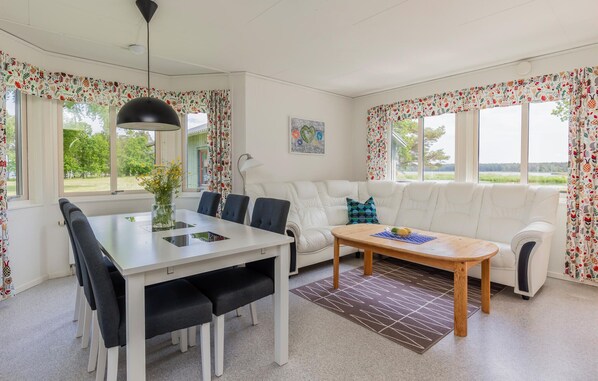 A modern living room with a white sofa, a dining table with chairs, and a coffee table. There are large windows with floral curtains, a hanging light fixture, and a painting on the wall.