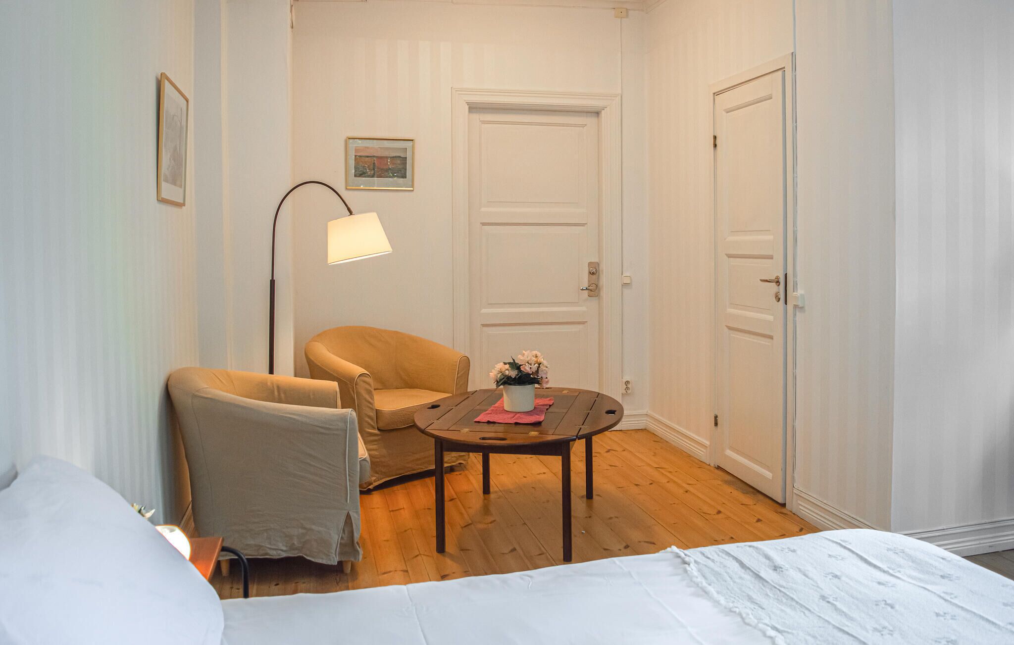 A room with a bed, two chairs, a small table with a vase, a floor lamp, and two doors.