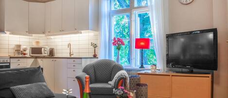 Un salon moderne comprenant un canapé, une table basse, un téléviseur et un coin cuisine avec des meubles blancs et un four à micro-ondes.