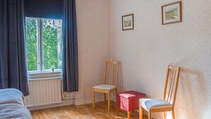 Una habitación con piso de madera, un radiador, dos sillas, una mesita y una ventana con cortinas.