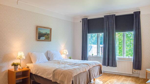 Un dormitorio con una cama grande, dos lámparas de noche, una mesita de noche, una ventana con cortinas y un cuadro en la pared.