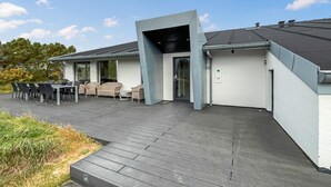 A modern outdoor patio area with a wooden deck, outdoor seating, and a glass door entrance. - Nice home in Blåvand with sauna (Blåvand)