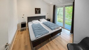 A modern bedroom with a bed, bedside tables, a headboard, a window with curtains, and a view of greenery outside. - Nice home in Blåvand with sauna (Blåvand)