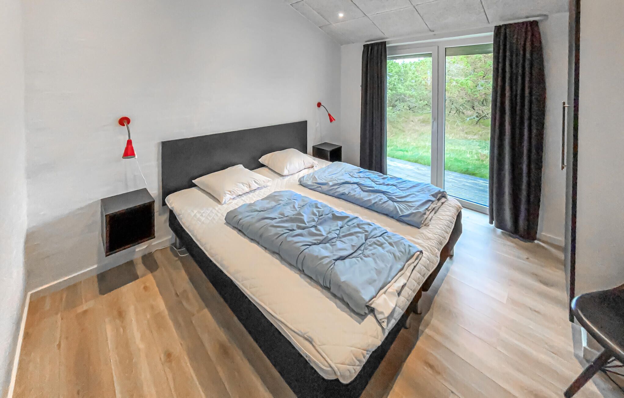 A modern bedroom with a large bed, two bedside tables, a red lamp, and a view of greenery through the window.