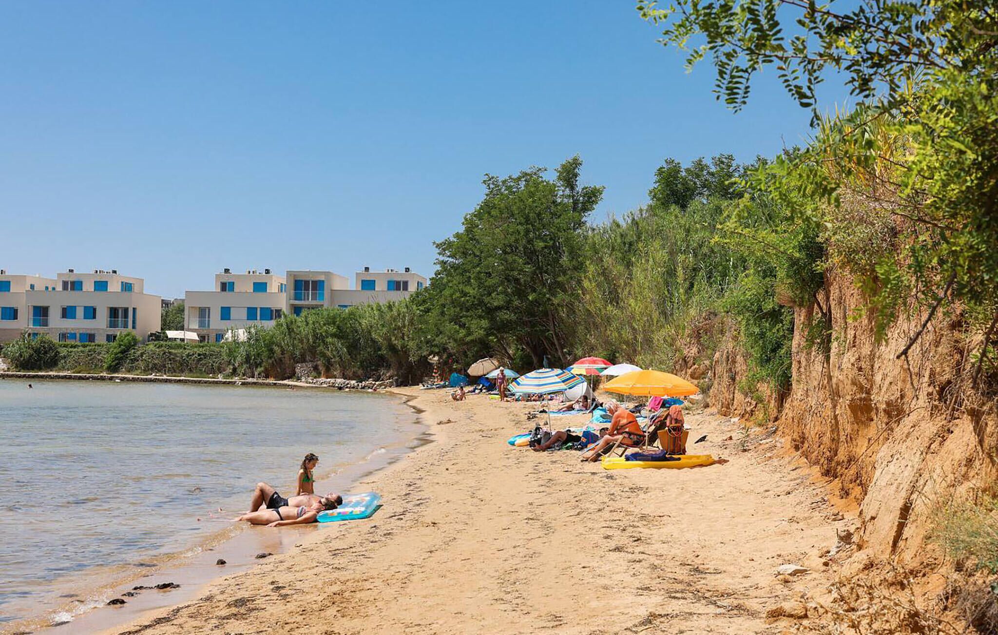 A beach with people sunbathing and swimming, a row of colorful umbrellas, and buildings in the background.
