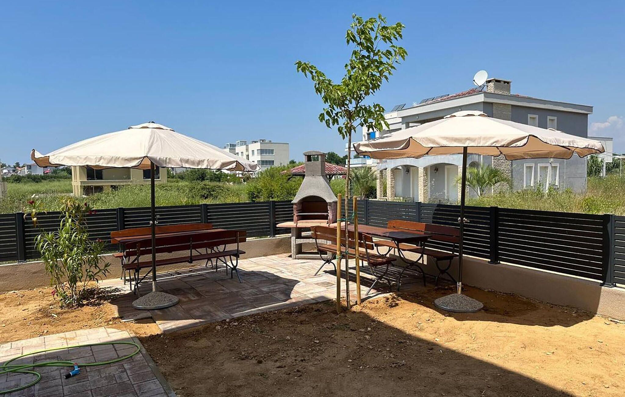 An outdoor patio with wooden tables and chairs, a barbecue grill, and umbrellas.