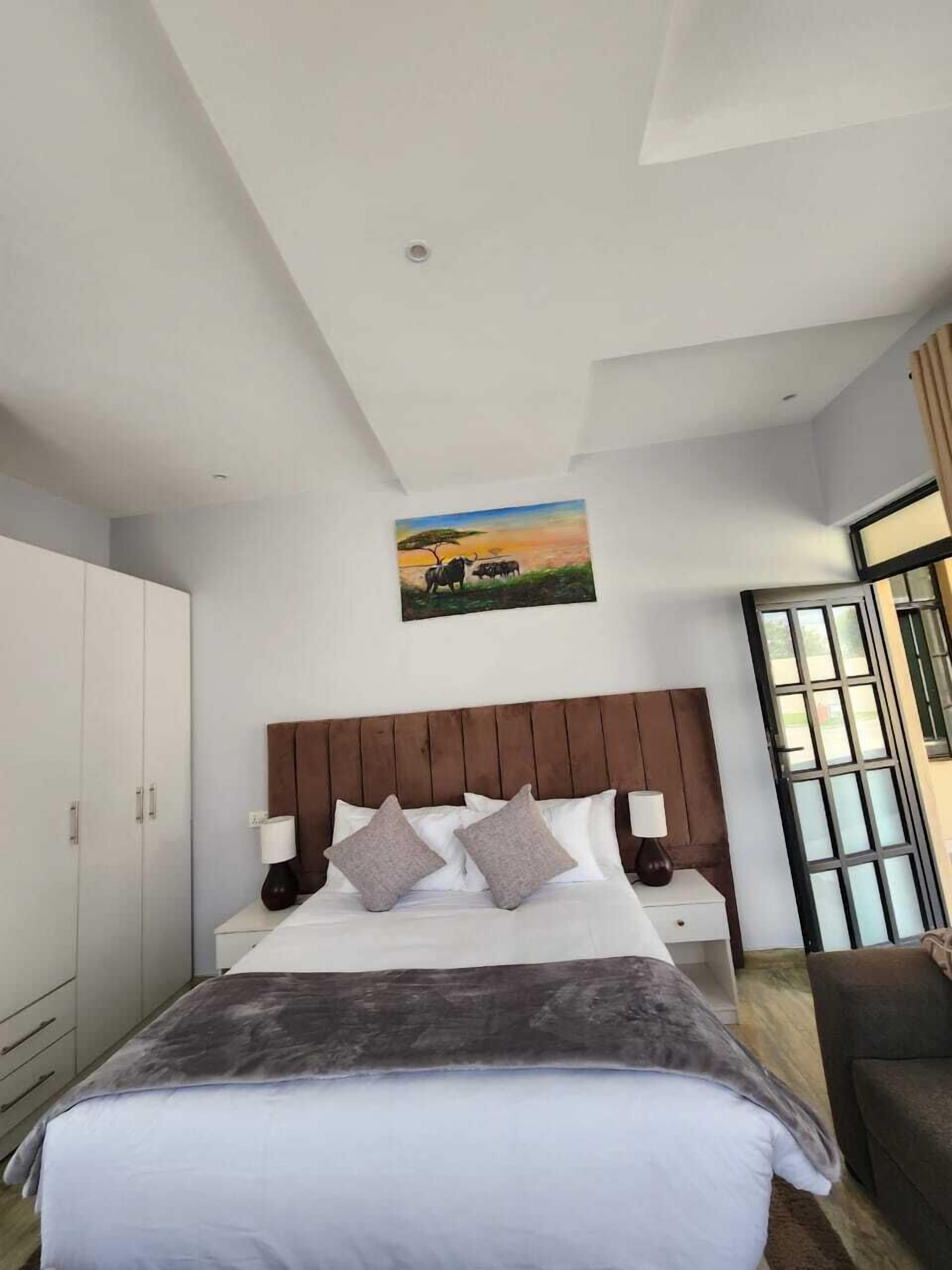 Quarto moderno com cama grande, cabeceira, mesa de cabeceira, abajur e pintura de uma paisagem de savana.
