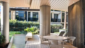 An outdoor patio area with a round table and chairs, surrounded by greenery and a view of the building's interior. - Elegant 1-bed Apartment With Premium Amenities (Melbourne)