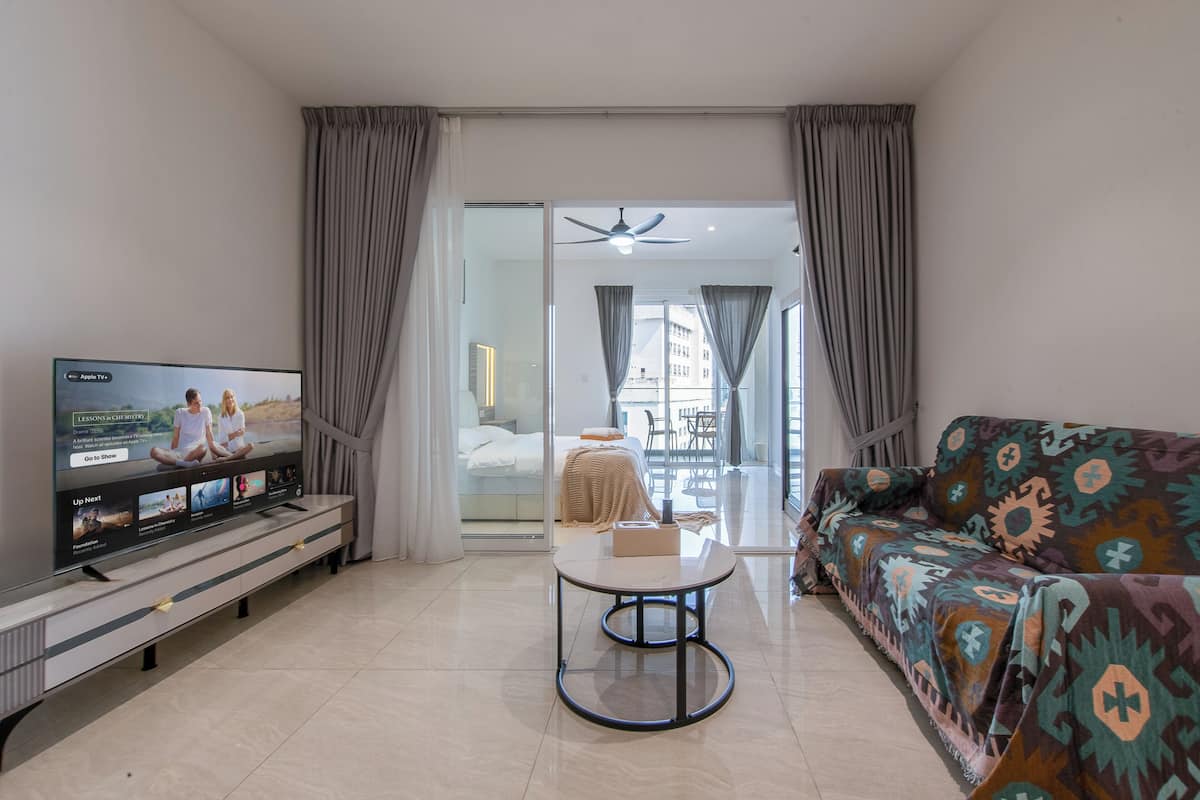 A modern hotel room with a TV, a sofa, a small table, and a sliding glass door leading to a balcony.