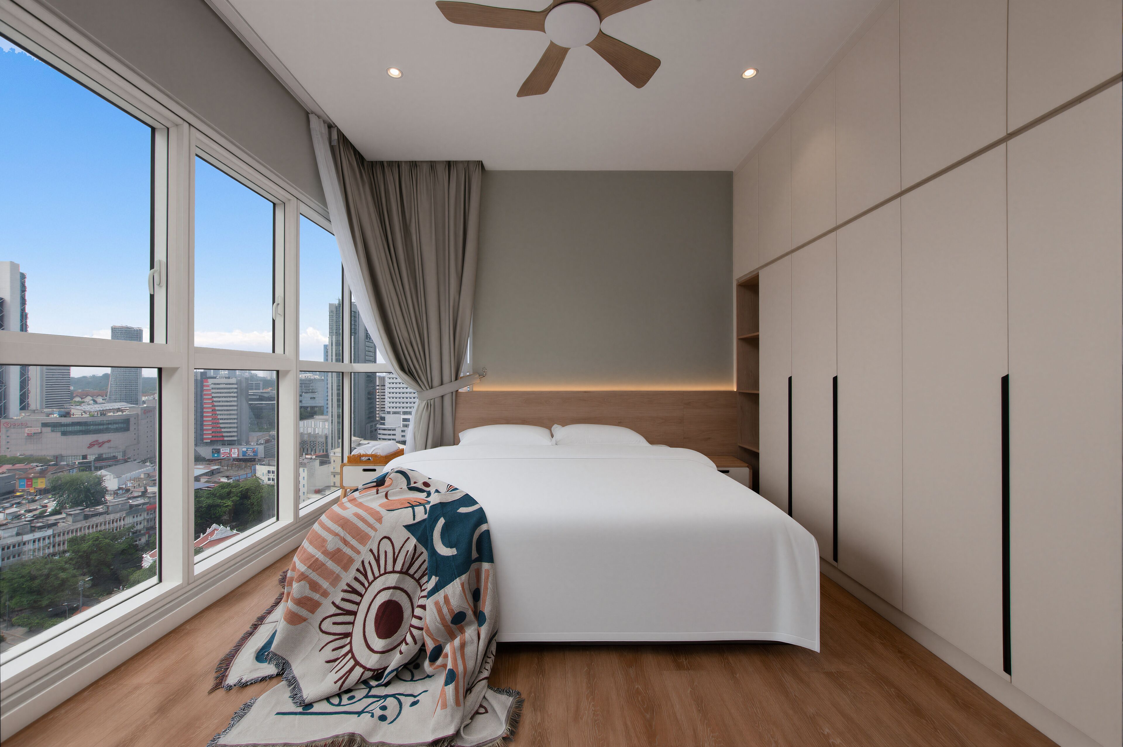 A modern bedroom with a large window, a bed, a ceiling fan, and a city view.