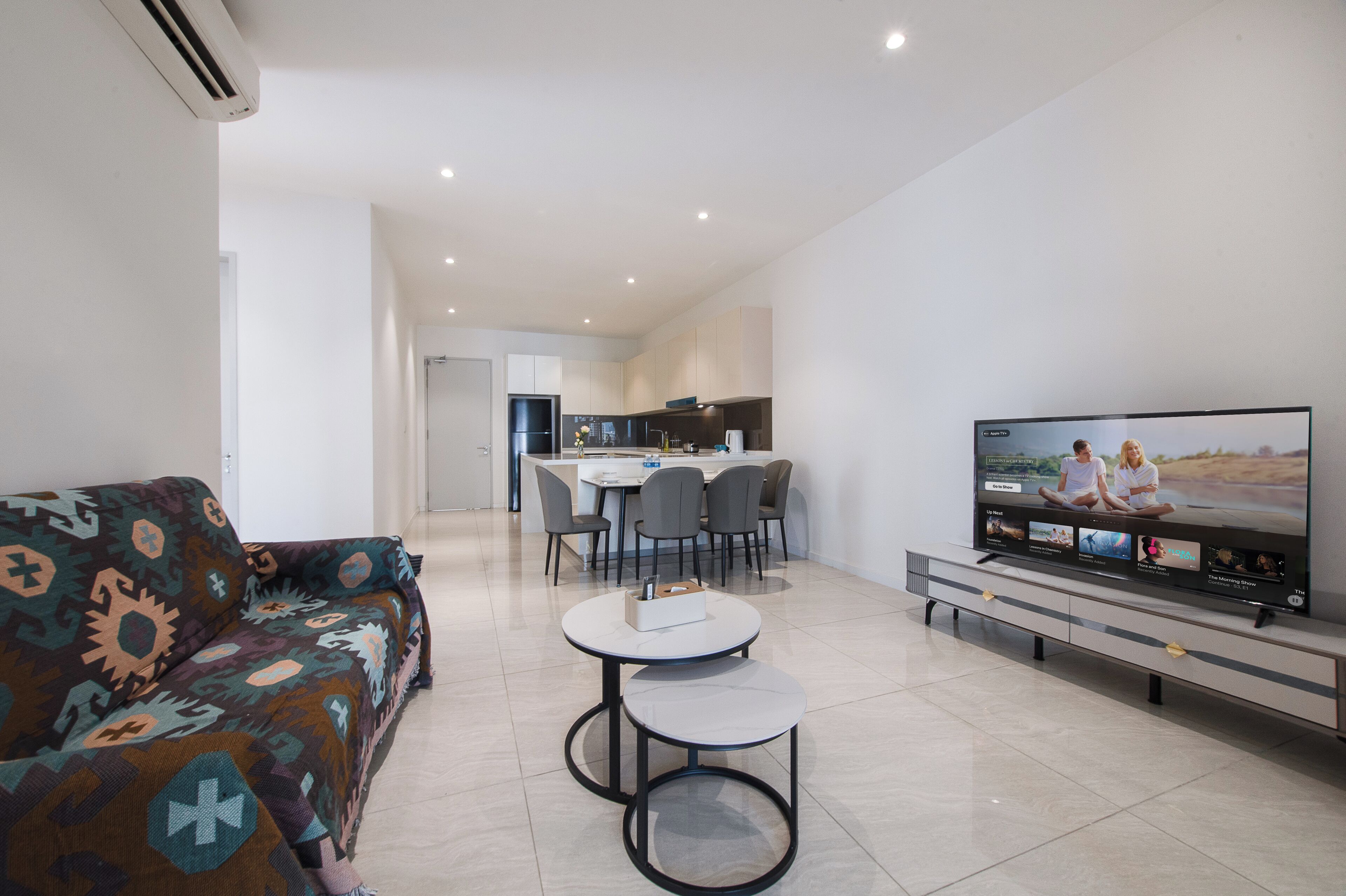 A modern living room with a sofa, two small tables, a TV, and a kitchen in the background.