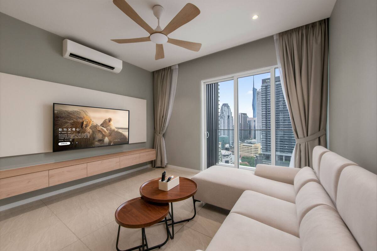 A modern living room with a flat-screen TV, a ceiling fan, a sofa, a coffee table, and a large window with a city view.